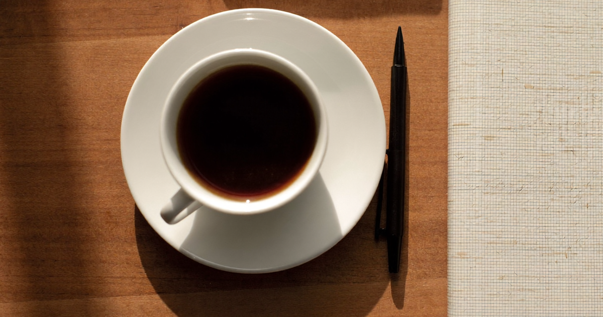Xícara branca de café preto vista de cima sobre mesa de madeira, com uma caneta preta e a borda de um caderno de capa clara ao lado, sob luz de janela
